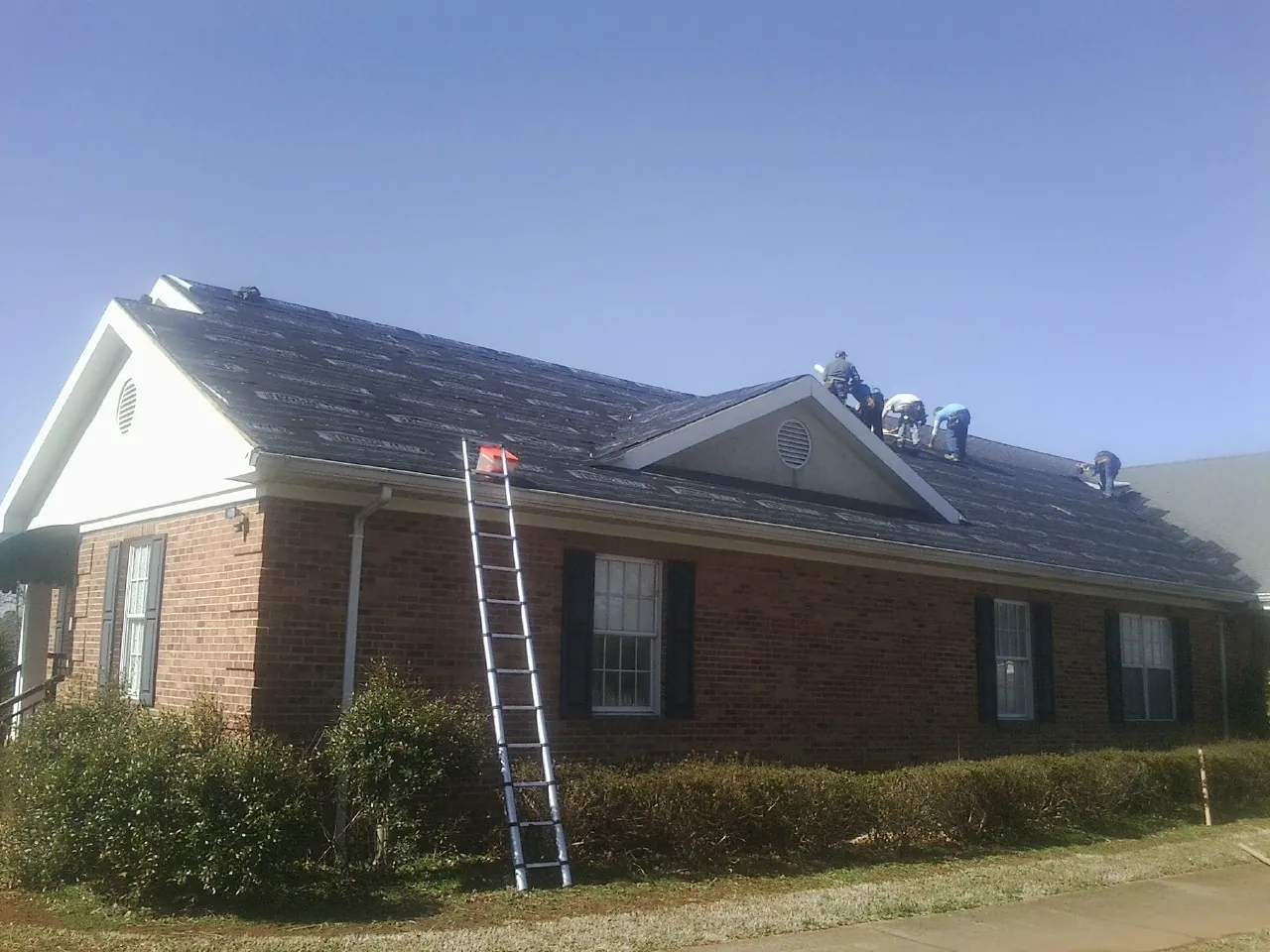 Crew tearing shingles off a brick ranch roof with a ladder set — Metal Roof Repair work by our crew in Trimble